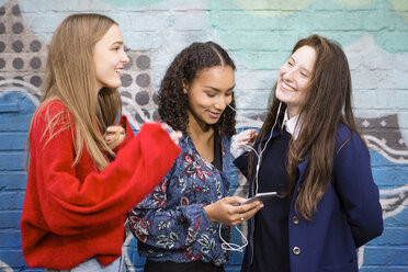 Three friends standing in front of graffiti wall hearing music with smartphone and earphones - OJF00237