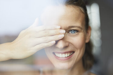 Porträt einer lachenden jungen Frau hinter einer Fensterscheibe, die ihr Auge verdeckt - PNEF00409