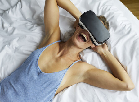 Lachende junge Frau im Bett liegend mit VR-Brille - PNEF00417