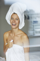 Portrait of happy young woman behind windowpane wrapped in towels - PNEF00419