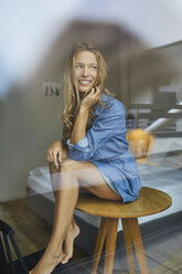 Lächelnde junge Frau sitzt auf einem Tisch hinter einer Fensterscheibe - PNEF00420