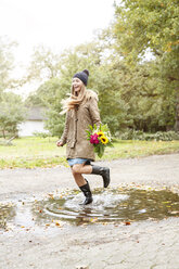 Glückliche junge Frau, die einen Blumenstrauß hält und in eine Pfütze springt - PESF00905