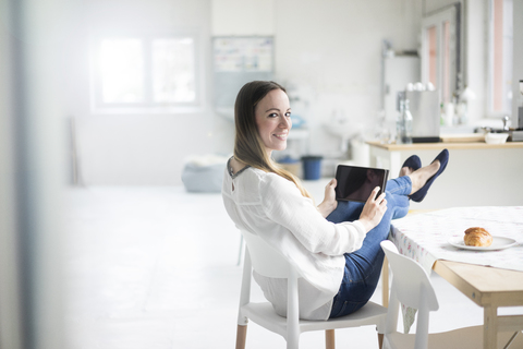 Porträt einer lächelnden Frau mit Tablet, die sich am Frühstückstisch in einem Loft entspannt, lizenzfreies Stockfoto