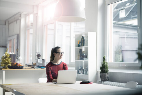 Lächelnde Frau sitzt am Tisch mit Laptop in einem Loft und schaut aus dem Fenster - MOEF00711