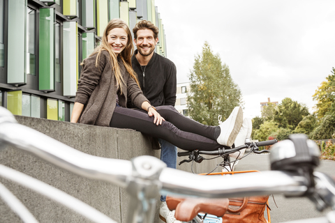 Porträt eines lächelnden Paares mit Fahrrädern, das an einer Wand sitzt, lizenzfreies Stockfoto