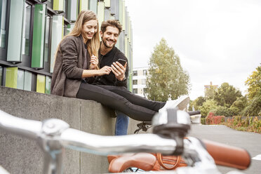 Glückliches Paar mit Fahrrädern, das auf einer Mauer sitzt und auf ein Mobiltelefon schaut - PESF00926