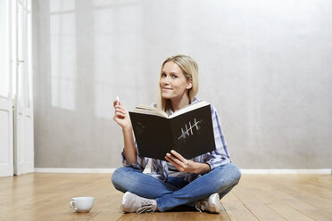 Portrait of smiling blond woman with book sitting on the floor - FMKF04746
