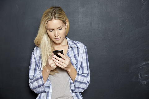 Blonde Frau steht vor einer Tafel und schaut auf ihr Handy, lizenzfreies Stockfoto