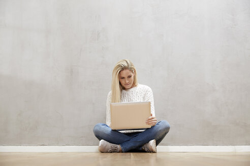 Blonde Frau sitzt auf dem Boden vor einer grauen Wand und benutzt einen Laptop - FMKF04751