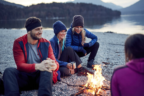 Smiling friends sitting around campfire at lakeshore - PNEF00433