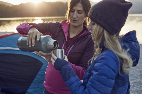 Two women with hot drink camping at lakeshore - PNEF00505