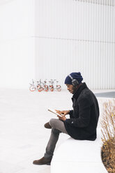 Italy, Milan, African american with headphone reading a book - MAUF01283