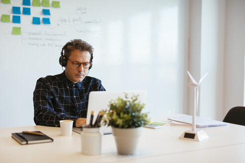 Geschäftsmann mit Kopfhörern und Laptop am Schreibtisch im Büro - EBSF02076