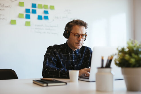 Geschäftsmann mit Kopfhörern und Laptop am Schreibtisch im Büro - EBSF02078