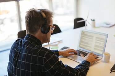 Geschäftsmann mit Kopfhörern und Laptop am Schreibtisch im Büro - EBSF02079