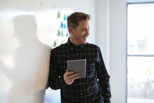Lächelnder Geschäftsmann mit Tablet im Büro lehnt gegen Whiteboard - EBSF02084