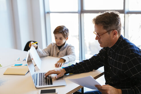 Geschäftsmann mit Laptop am Schreibtisch im Büro mit Sohn sitzt neben ihm - EBSF02096