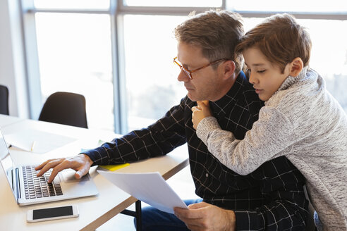 Businessman using laptop at desk in office with son embracing him - EBSF02097