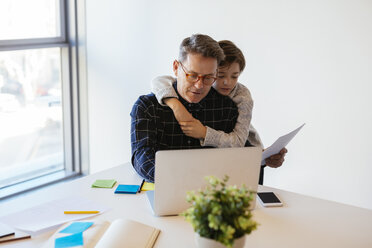 Businessman using laptop at desk in office with son embracing him - EBSF02098