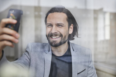 Porträt eines lachenden Mannes hinter einer Fensterscheibe, der ein Selfie mit seinem Mobiltelefon macht - PNEF00513