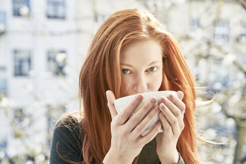 Porträt einer rothaarigen Frau, die auf einem Balkon eine Schale mit weißem Kaffee trinkt - FMKF04764