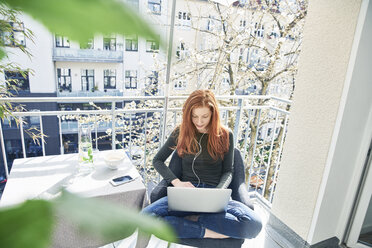 Redheaded woman sitting on balcony using laptop and earphones in spring - FMKF04765