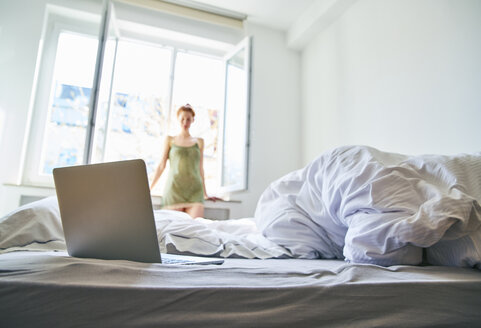 Laptop auf dem Bett mit einer Frau, die vor einem Fenster im Hintergrund steht - FMKF04769