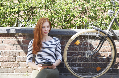 Portrait of redheaded woman with tablet and bicycle sitting on a wall stock photo