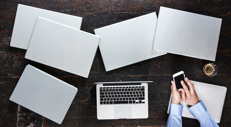 Man's hands using smartphone on table with seven laptops, top view - FMKF04786