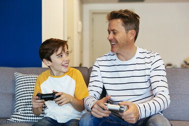 Happy father and son playing video game on couch at home - EBSF02112