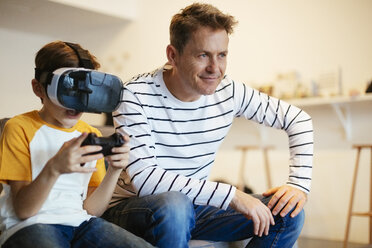 Boy wearing VR glasses playing video game with father on couch at home - EBSF02116