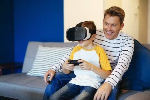Junge mit VR-Brille spielt Videospiel mit Vater auf der Couch zu Hause, lizenzfreies Stockfoto