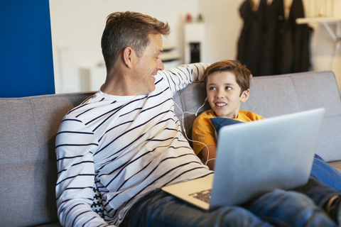 Lächelnder Vater und Sohn mit Ohrstöpseln und Laptop auf der Couch zu Hause, lizenzfreies Stockfoto