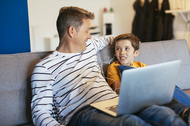 Lächelnder Vater und Sohn mit Ohrstöpseln und Laptop auf der Couch zu Hause - EBSF02141