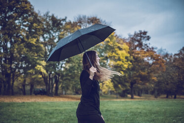 Glückliche junge Frau mit Regenschirm, die in einem herbstlichen Park spazieren geht - JSCF00029