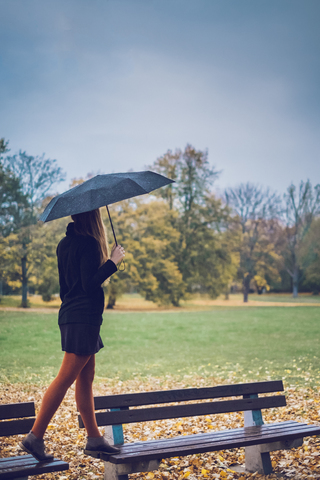 Junge Frau mit Regenschirm balanciert auf zwei Bänken in einem herbstlichen Park, lizenzfreies Stockfoto