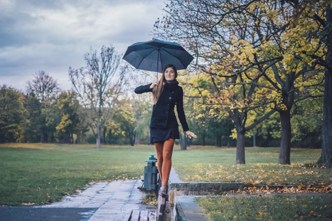 Junge Frau mit Regenschirm balanciert auf der Rückenlehne einer Bank in einem herbstlichen Park, lizenzfreies Stockfoto