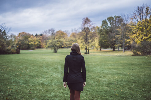 Rückenansicht einer jungen Frau, die auf einer Wiese in einem herbstlichen Park spazieren geht - JSCF00047