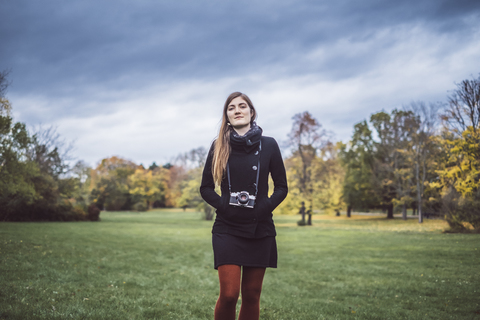 Porträt einer jungen Frau mit Kamera, die auf einer Wiese in einem herbstlichen Park spazieren geht, lizenzfreies Stockfoto