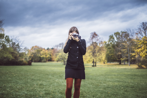 Junge Frau fotografiert mit Kamera auf einer Wiese im herbstlichen Park, lizenzfreies Stockfoto