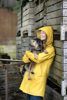Lächelnde Frau auf einem Bauernhof, die an Holzkisten steht und einen Hund hält - PESF00956