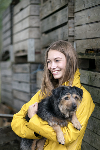 Lächelnde Frau auf einem Bauernhof, die an Holzkisten steht und einen Hund hält, lizenzfreies Stockfoto