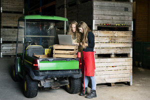 Zwei Frauen benutzen einen Laptop zwischen Kisten auf einem Bauernhof - PESF00980