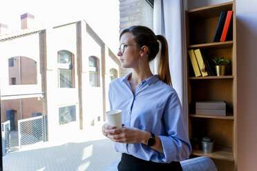 Young woman with coffee cup looking out of office window - VABF01462