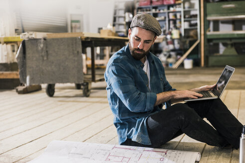 Man with laptop sitting on the floor looking at construction plan - UUF12694