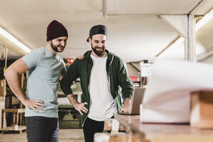 Zwei lächelnde junge Männer vor einem Laptop in einer Werkstatt - UUF12707