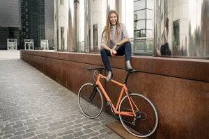 Glückliche junge Frau mit Fahrrad, die eine Pause in der Stadt macht und einen Apfel isst - PESF01000