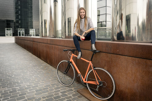 Happy young woman with bicycle having a break in the city eating an apple - PESF01000