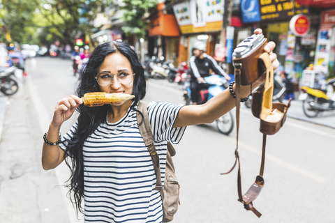 Vietnam, Hanoi, junge Frau macht ein Selfie mit altmodischer Kamera auf der Straße und isst einen Maiskolben, lizenzfreies Stockfoto