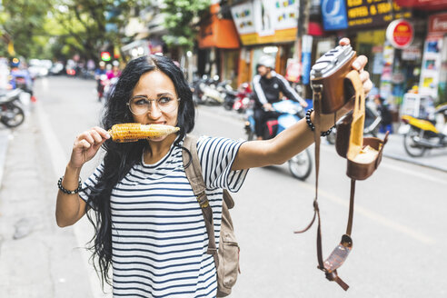 Vietnam, Hanoi, young woman taking a selfie with old-fashioned camera on the street eating a corn cob - WPEF00063